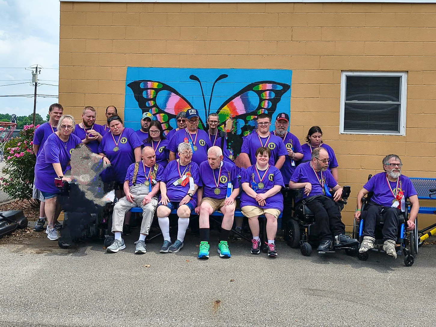 A large group of adults wearing medals and purple Special Olympics shirts, smiling in front of a rainbow butterfly mural.